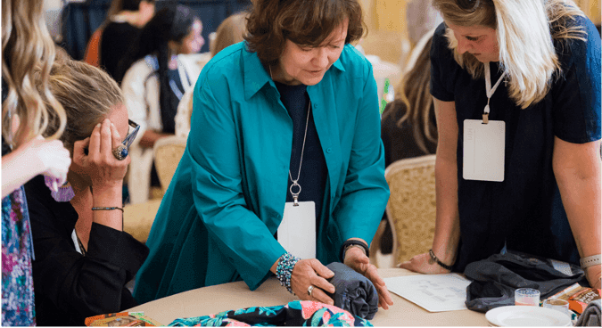A woman is practicing folding at a round-table of course attendees, who are watching her