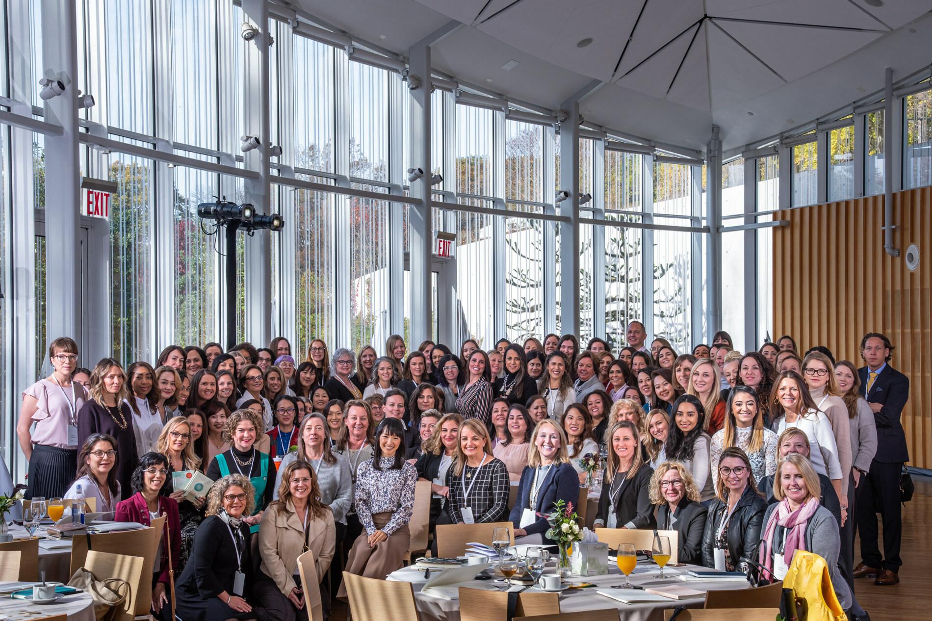 A large group of over 100 diverse attendees of the KonMari Consultant Course at a spacious venue with large windows and natural light with Marie Kondo sitting in the middle